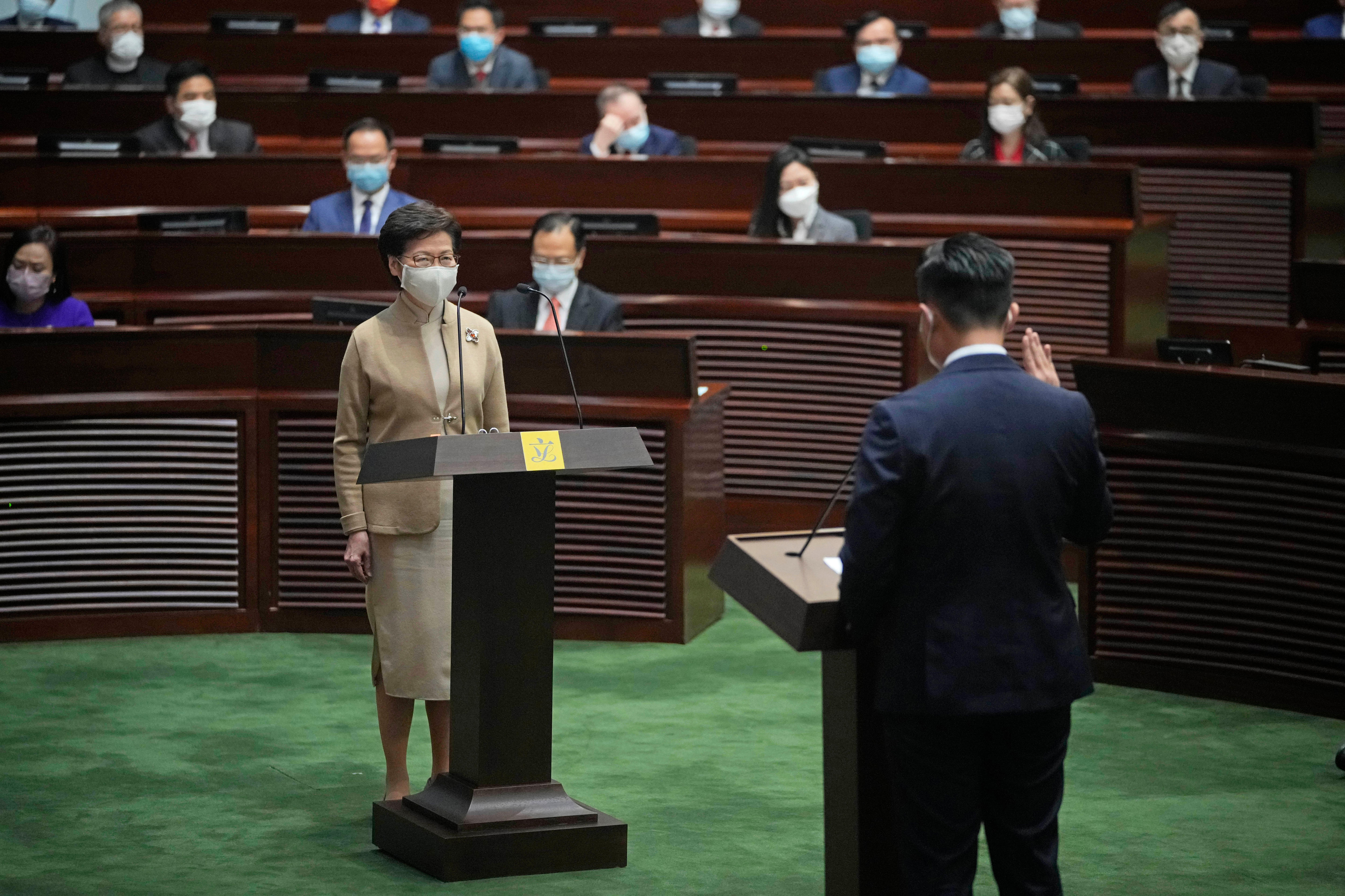 A man wearing a suit stands in front of Carrie Lam and raises his hand in oath.